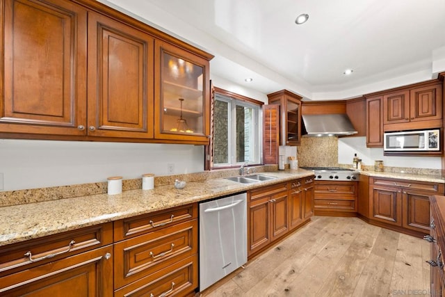 kitchen with light stone counters, wall chimney exhaust hood, stainless steel appliances, sink, and light hardwood / wood-style floors