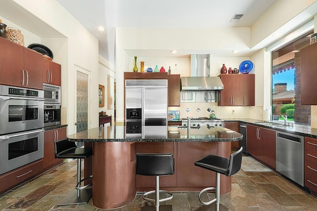 kitchen with a kitchen island with sink, wall chimney range hood, built in appliances, decorative backsplash, and a breakfast bar area