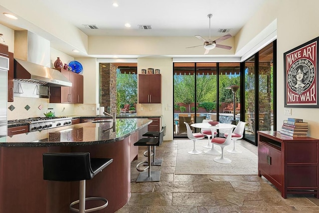 kitchen with ceiling fan, wall chimney range hood, tasteful backsplash, an island with sink, and a breakfast bar