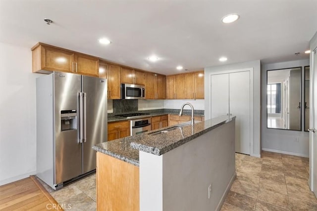 kitchen with an island with sink, stainless steel appliances, decorative backsplash, dark stone countertops, and sink