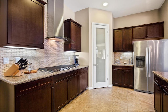 kitchen with dark brown cabinetry, wall chimney exhaust hood, stainless steel appliances, light stone counters, and backsplash