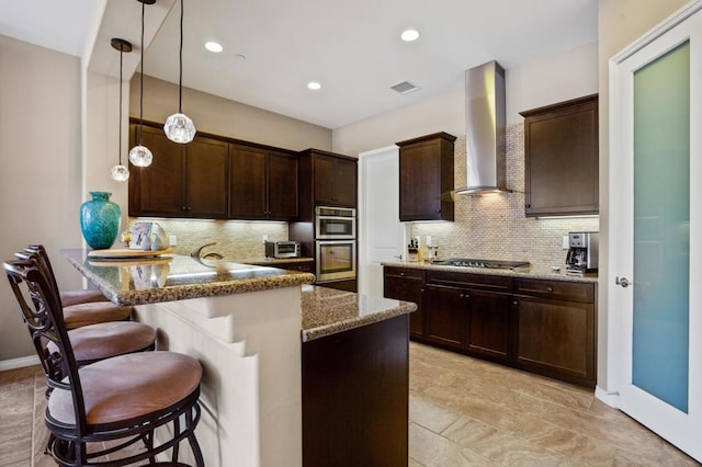 kitchen featuring kitchen peninsula, wall chimney exhaust hood, stainless steel appliances, decorative light fixtures, and a breakfast bar area