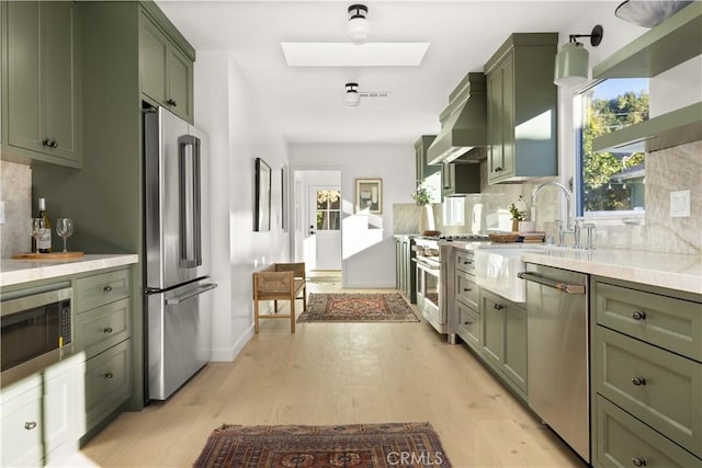 kitchen featuring backsplash, wall chimney range hood, a skylight, light wood-type flooring, and stainless steel appliances