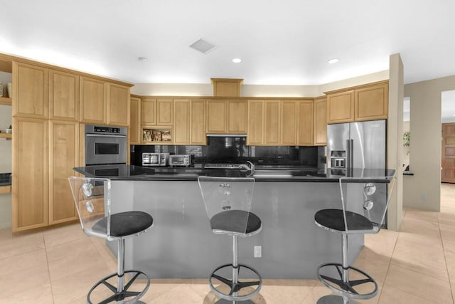 kitchen featuring tasteful backsplash, a kitchen bar, a kitchen island with sink, stainless steel appliances, and light tile patterned floors