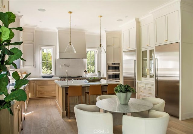 kitchen with crown molding, light hardwood / wood-style flooring, an island with sink, appliances with stainless steel finishes, and decorative light fixtures