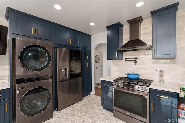 kitchen featuring stacked washer and dryer, stainless steel appliances, blue cabinets, and wall chimney range hood