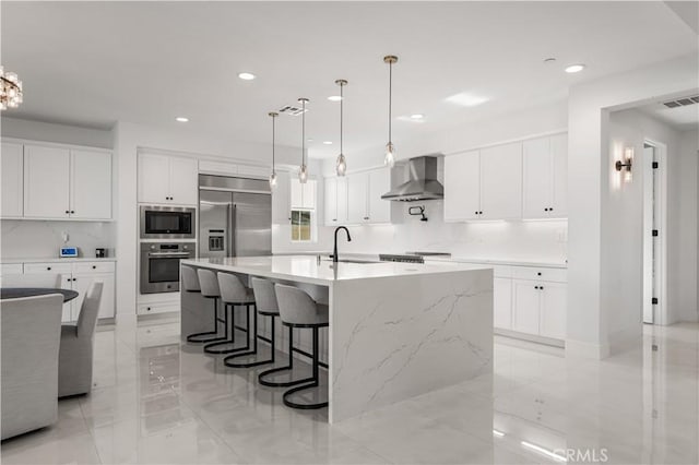 kitchen with built in appliances, an island with sink, hanging light fixtures, and wall chimney range hood