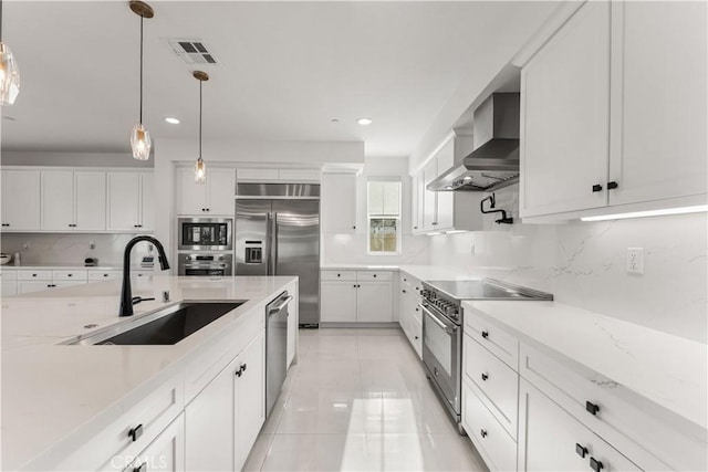 kitchen with pendant lighting, wall chimney range hood, sink, built in appliances, and white cabinetry