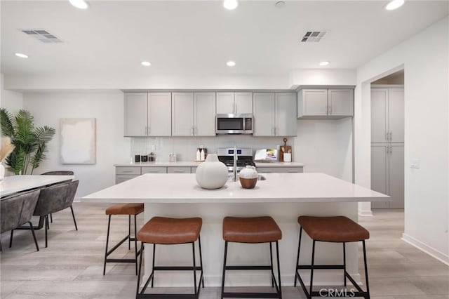 kitchen featuring tasteful backsplash, gray cabinets, light wood-type flooring, an island with sink, and stainless steel appliances