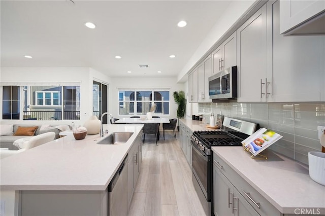 kitchen with light hardwood / wood-style floors, sink, appliances with stainless steel finishes, an island with sink, and gray cabinetry