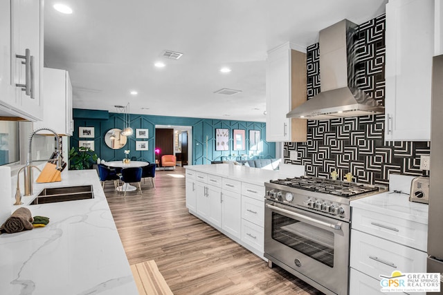 kitchen featuring wall chimney range hood, sink, high end stainless steel range, light wood-type flooring, and white cabinetry