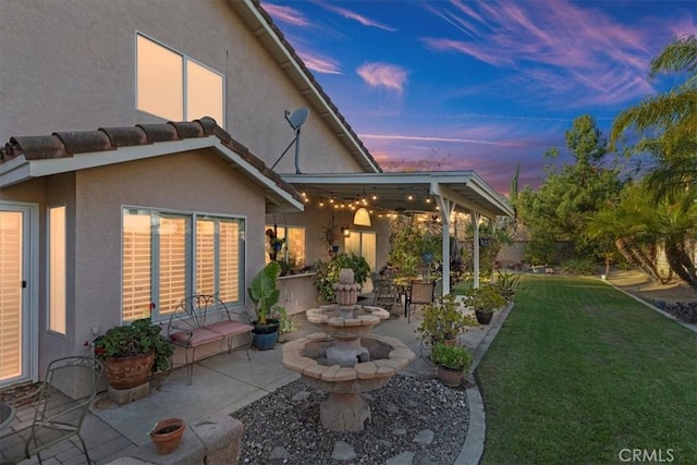 back house at dusk featuring a lawn and a patio area