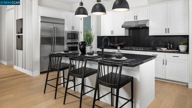 kitchen with built in appliances, decorative light fixtures, tasteful backsplash, light hardwood / wood-style floors, and a kitchen island with sink