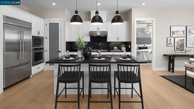 kitchen featuring built in appliances, an island with sink, decorative backsplash, and white cabinetry