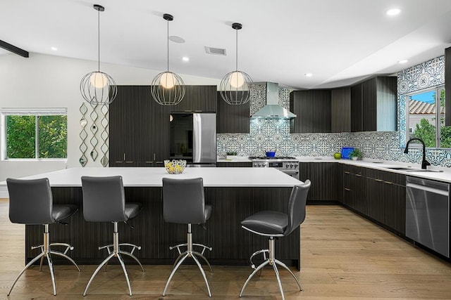 kitchen with sink, stainless steel appliances, wall chimney range hood, light hardwood / wood-style flooring, and a kitchen island