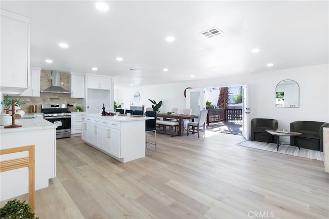 kitchen with white cabinets, wall chimney range hood, light hardwood / wood-style flooring, stainless steel range, and a kitchen island