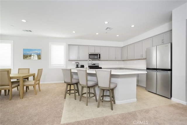 kitchen with appliances with stainless steel finishes, a breakfast bar, light colored carpet, a kitchen island with sink, and gray cabinets