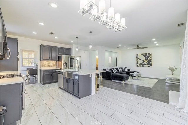 kitchen with appliances with stainless steel finishes, ceiling fan with notable chandelier, a kitchen island with sink, sink, and hanging light fixtures