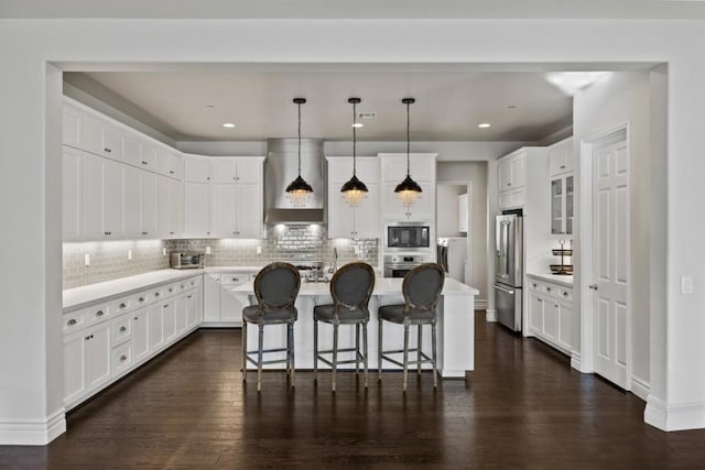 kitchen with hanging light fixtures, an island with sink, stainless steel appliances, white cabinets, and wall chimney exhaust hood