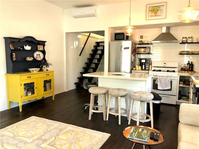 kitchen featuring hanging light fixtures, a wall mounted air conditioner, wall chimney range hood, a kitchen breakfast bar, and appliances with stainless steel finishes