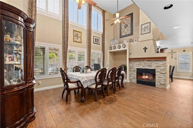 dining room featuring a stone fireplace, ceiling fan, hardwood / wood-style floors, and a towering ceiling