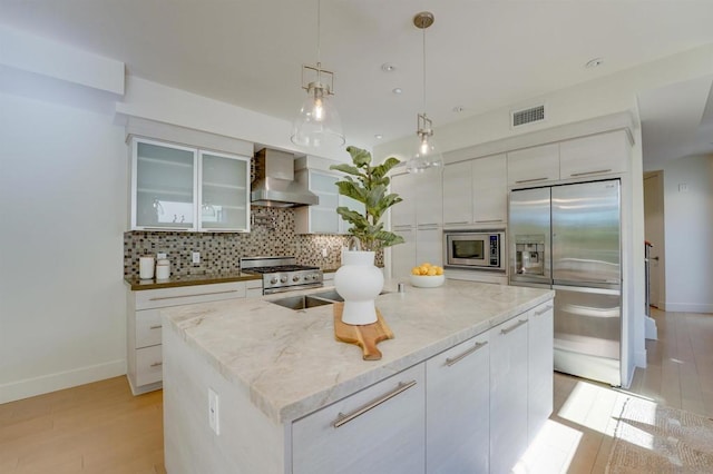 kitchen with wall chimney exhaust hood, light wood-type flooring, an island with sink, white cabinetry, and stainless steel appliances