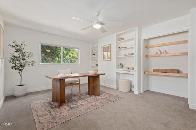 unfurnished office featuring built in shelves, baseboards, a ceiling fan, and light colored carpet