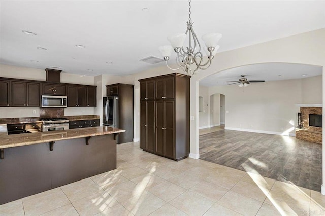 kitchen featuring dark brown cabinetry, stainless steel appliances, a kitchen breakfast bar, decorative light fixtures, and ceiling fan with notable chandelier