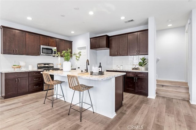 kitchen featuring a kitchen island with sink, a kitchen breakfast bar, appliances with stainless steel finishes, dark brown cabinets, and light hardwood / wood-style floors