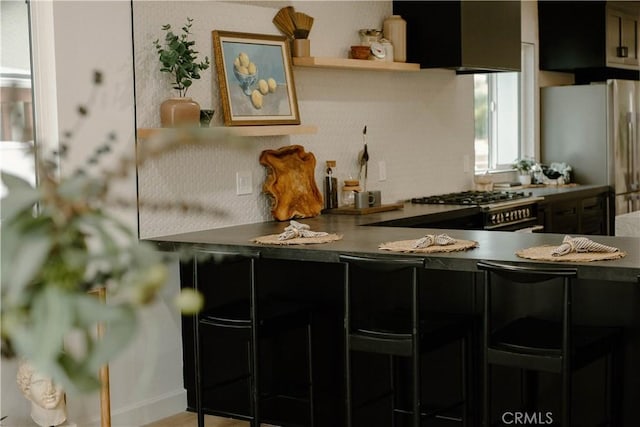 kitchen featuring open shelves, freestanding refrigerator, wall chimney exhaust hood, range with gas stovetop, and a kitchen bar