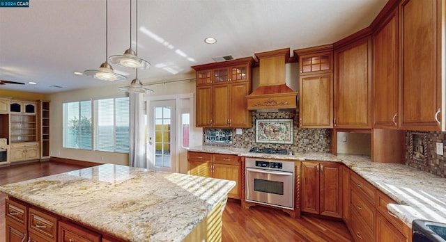 kitchen with appliances with stainless steel finishes, premium range hood, light stone counters, a center island, and hanging light fixtures