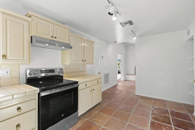 kitchen with stainless steel electric range oven, tile counters, rail lighting, backsplash, and cream cabinets