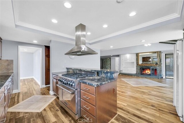 kitchen with dark stone counters, island range hood, stainless steel stove, a fireplace, and light hardwood / wood-style floors