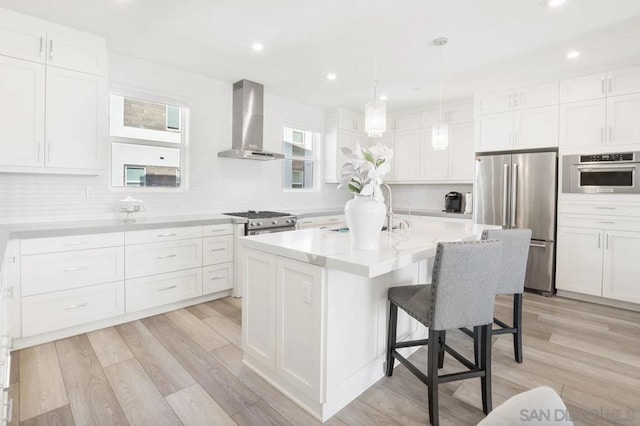 kitchen featuring a center island, wall chimney range hood, appliances with stainless steel finishes, white cabinets, and light wood-type flooring