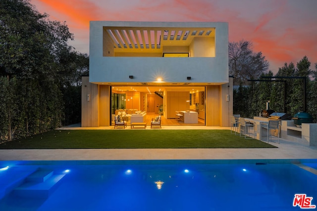 back house at dusk featuring exterior kitchen, a patio, an outdoor hangout area, and a lawn