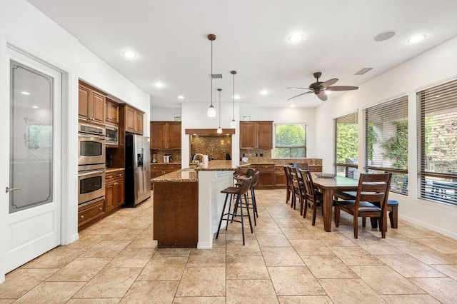 kitchen featuring hanging light fixtures, ceiling fan, an island with sink, appliances with stainless steel finishes, and tasteful backsplash