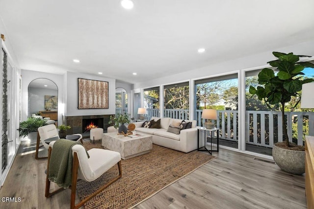 living area with arched walkways, light wood-type flooring, a tile fireplace, and recessed lighting