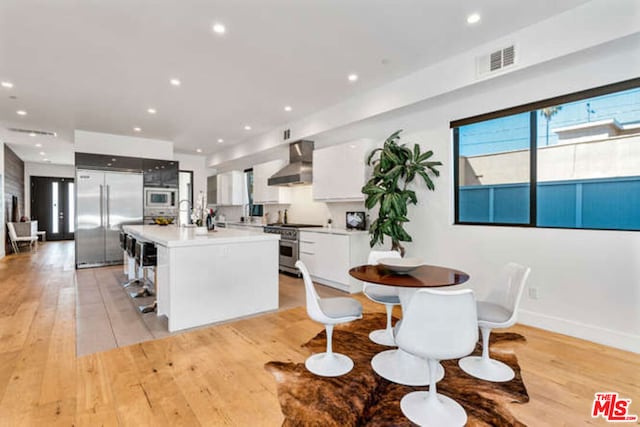 kitchen with a center island with sink, wall chimney range hood, built in appliances, light wood-type flooring, and white cabinetry