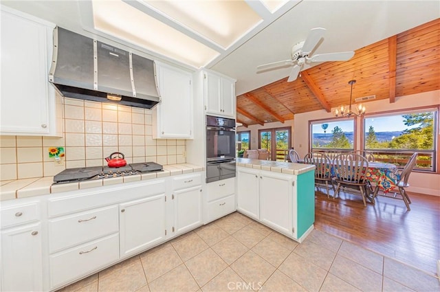 kitchen featuring a warming drawer, tile counters, wall chimney range hood, black oven, and stovetop