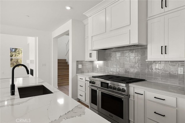 kitchen with range with two ovens, backsplash, white cabinetry, a sink, and light stone countertops