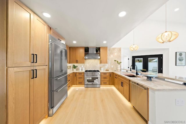 kitchen with light wood-type flooring, premium appliances, sink, wall chimney range hood, and hanging light fixtures