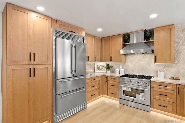 kitchen featuring backsplash, light stone counters, wall chimney exhaust hood, light hardwood / wood-style flooring, and high quality appliances