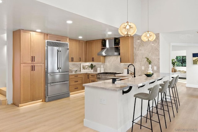 kitchen with wall chimney exhaust hood, light wood-type flooring, light brown cabinetry, kitchen peninsula, and stainless steel appliances