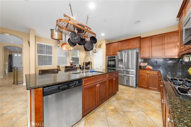 kitchen featuring a kitchen island with sink, sink, decorative backsplash, dark stone countertops, and appliances with stainless steel finishes