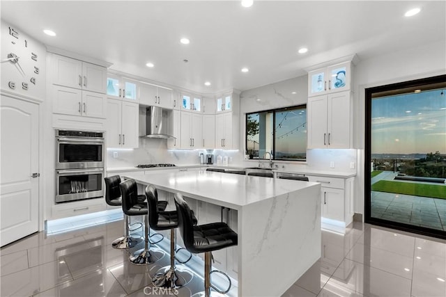 kitchen with wall chimney exhaust hood, a kitchen bar, white cabinetry, double oven, and a kitchen island