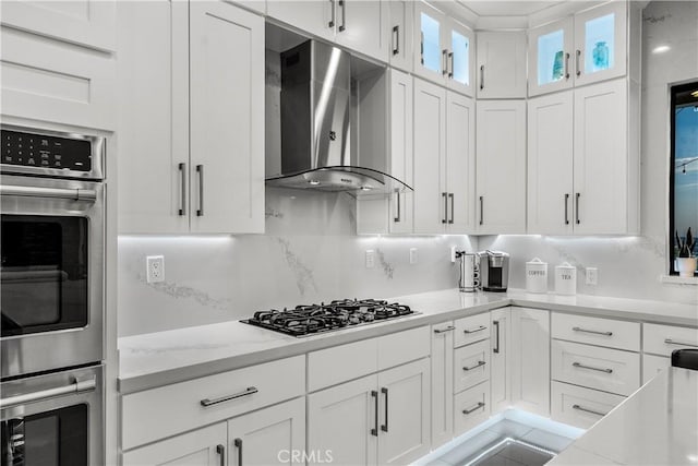 kitchen featuring appliances with stainless steel finishes, white cabinetry, light stone counters, decorative backsplash, and wall chimney exhaust hood