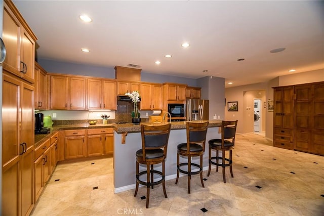 kitchen with black microwave, an island with sink, a breakfast bar area, dark stone counters, and stainless steel fridge with ice dispenser