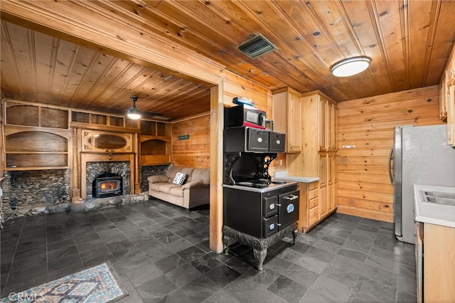 kitchen with light brown cabinets, wooden ceiling, a wood stove, stainless steel refrigerator, and wood walls