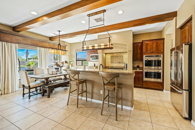 kitchen with beam ceiling, hanging light fixtures, light stone counters, a kitchen island with sink, and appliances with stainless steel finishes