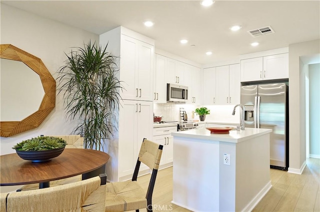 kitchen featuring tasteful backsplash, appliances with stainless steel finishes, a kitchen island with sink, white cabinets, and light wood-type flooring